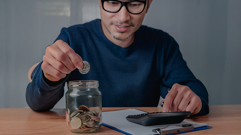 Man dropping money in savings jar with notepad and calculator on his desk